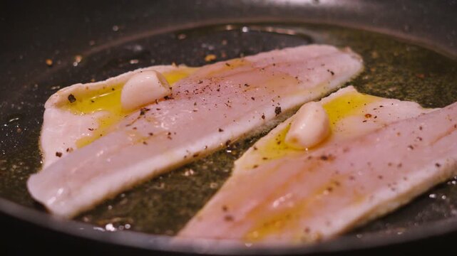 Rotating view of two sea bass fillets in a hot pan cooking skin side down in bubbling olive oil. White fish cooked with garlic, salt, pepper. Healthy omega-3 rich, high protein food. 4K.