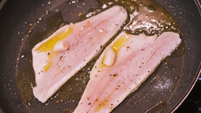 Top down of two sea bass fillets in a hot pan cooking skin side down in bubbling olive oil. White fish cooked with garlic. Healthy omega-3 rich, high protein food. 4K.