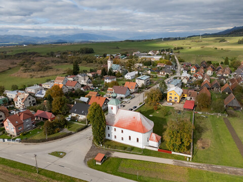 Aerial view of the white church with a red roof stands out amidst the rustic houses, bordered by fields and distant mountains, Haj, Zilina Region, Slovakia.