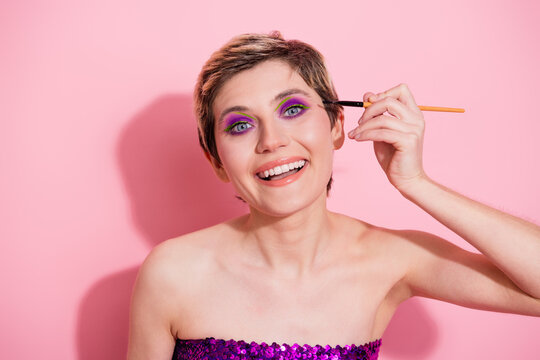 Playful glamour model applying bold colorful eye makeup with pink backdrop in a sequined purple top smiling at the camera for fashion marketing beauty lifestyle image