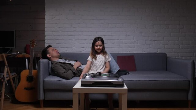 Father and daughter relax on a sofa, listening to vinyl on a vintage turntable, sharing a cozy, quiet evening of music and intergenerational bonding at home