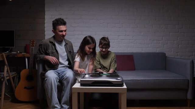 Father and two children relaxing on a sofa at home, enjoying music from a retro record player, sharing a special bonding moment during an evening together