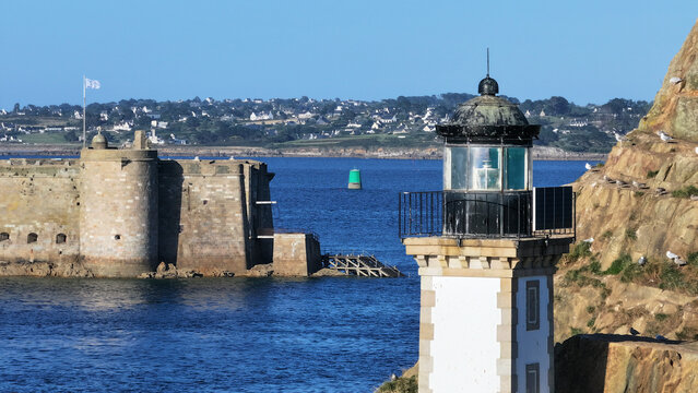 Aerial view of a white lighthouse on rugged cliffs, overlooking the deep blue sea and a formidable stone fort, Carantec, Brittany, France.