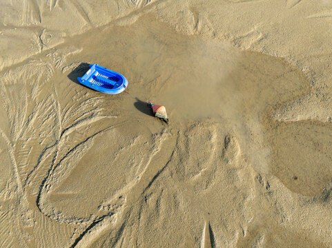 Aerial view of a child's blue plastic boat and a small cone resting on the patterned, sandy shore, Port Ostreicole de Bourcefranc-le-Chapus, Bourcefranc-le-Chapus, France.