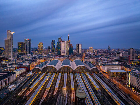 Aerial view of Hauptbahnhof's grand arches and tracks converging with the glowing modern city skyline at twilight, Frankfurt am Main, Hessen, Germany.