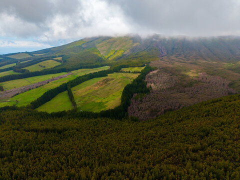 Aerial view of a dramatic landscape where vibrant green fields and lush forests meet desolate, scorched earth under a cloudy sky, Terceira, Azores, Portugal.
