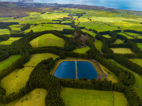 Aerial view of vibrant green pastures intricately bordered by dark emerald trees, encircling a tranquil sapphire reservoir under a clear sky, Terceira, Azores, Portugal.