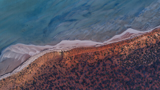 Aerial view of the dramatic contrast where rich red earth meets a pale sandy shore and the clear turquoise waters of the coast, Denham, Western Australia, Australia.