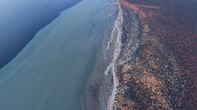 Aerial view of the vivid junction of deep blue ocean, turquoise bay, and a rugged reddish-brown arid coast, Denham, Western Australia, Australia.