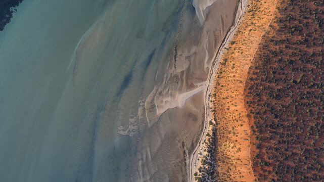 Aerial view of the stark boundary of blue-green water with sand ripples meeting vivid reddish-orange desert land, Denham, Western Australia, Australia.
