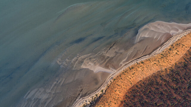 Aerial view of vivid red earth meeting the shallow, rippled turquoise waters, with intricate sand patterns visible beneath the surface, Denham, Western Australia, Australia.