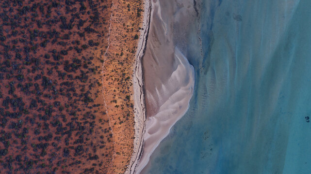 Aerial view of the vivid red earth meeting the calm, clear turquoise waters and rippled sandy seabed, Denham, Western Australia, Australia.