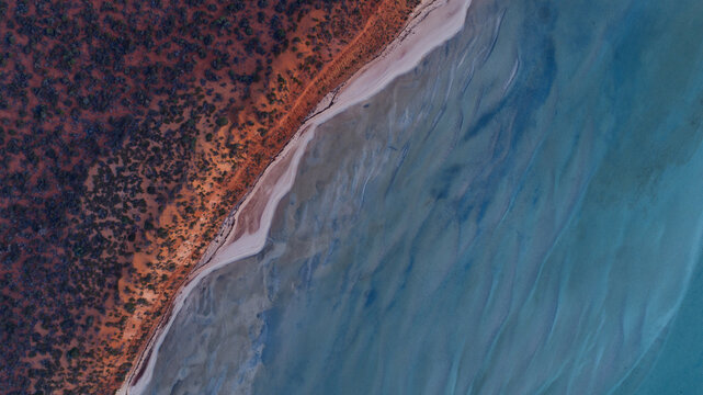 Aerial view of arid red earth meeting the serene, shallow blue-green waters of a calm bay along a pristine sandy shore, Denham, Western Australia, Australia.