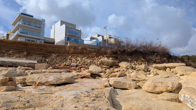 Wide shot of rugged eroded limestone coast in Malta with white modern seafront apartment blocks rising above sandy cliff, blue and grey cloudy sky