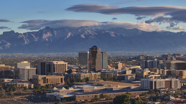 Aerial view of a vibrant cityscape bathed in warm light, contrasting with the rugged, majestic mountains under a dramatic sky, Tucson, Arizona, United States.