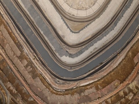 Aerial view of a vast, terraced industrial excavation with dark, winding water channels carving through contrasting strata of reddish-brown and gray earth, Tucson, Arizona, United States.