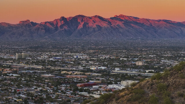 Aerial view of the rugged mountain range bathed in the fiery hues of sunset, casting long shadows over the sprawling city below, Tucson, Arizona, United States.