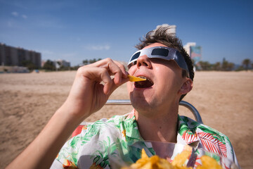 Naklejka premium Person wearing protective eclipse glasses on a sunny beach, relaxing and eating potato chips, balancing scientific interest with casual summer leisure during a celestial event