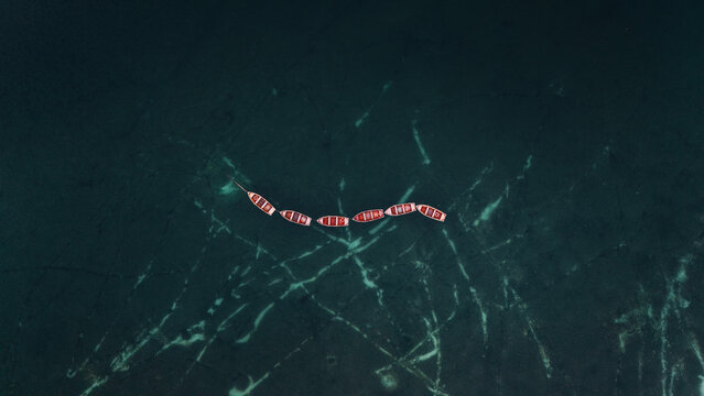 Aerial view of a striking line of crimson boats contrasting with the deep emerald water, etched with mysterious pale lines, Lago di Braies, Trentino-South Tyrol, Italy.