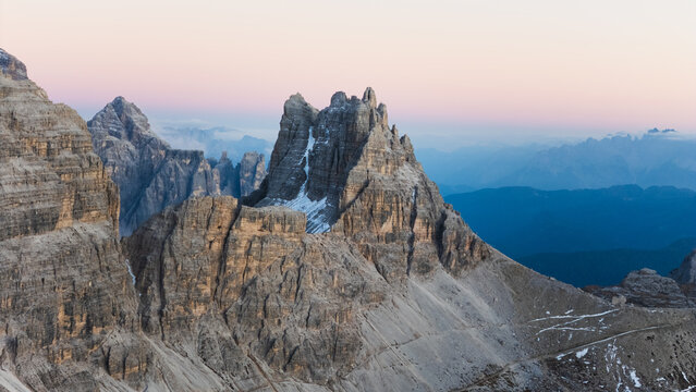 Aerial view of towering, rugged mountains with stark textures and lingering snow, illuminated by the gentle hues of a twilight sky, Tre Cime di Lavaredo, Veneto, Italy.