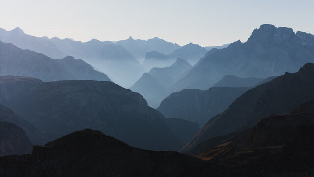 Aerial view of layered mountain ranges, dark foreground peaks contrasting soft, misty blue distant horizons, Tre Cime di Lavaredo, Veneto, Italy.