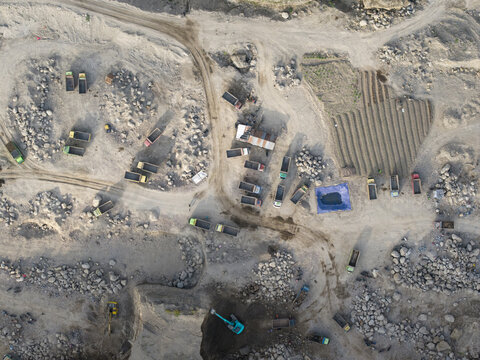Aerial view of a vast, rugged quarry site with numerous trucks and excavators amidst stark grey rock piles and disturbed earth, Klaten, Central Java, Indonesia.