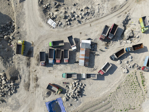 Aerial view of numerous colorful dump trucks scattered across a rugged, dusty industrial site with piles of raw materials under the harsh sun, Klaten, Central Java, Indonesia.