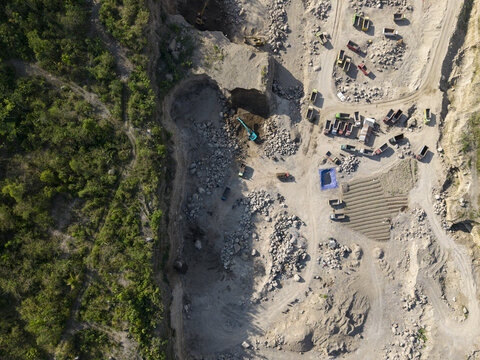 Aerial view of a stark contrast between a vibrant green forest and an active quarry with an excavator carving into the pale earth, Klaten, Central Java, Indonesia.