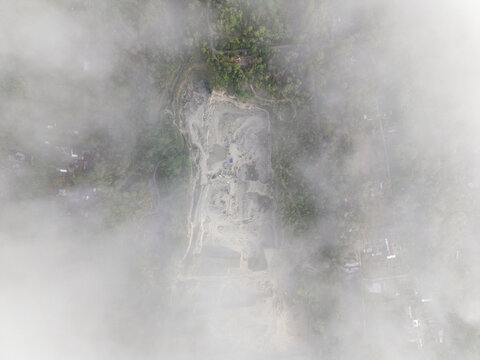 Aerial view of a pale quarry site through soft clouds, contrasting with lush green terrain and buildings, Klaten, Central Java, Indonesia.