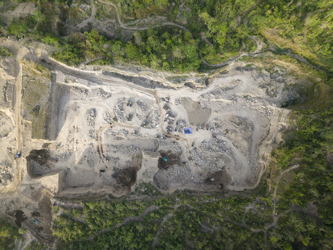 Aerial view of a massive open-pit quarry, its pale, excavated earth starkly contrasting with the vibrant green forest, Klaten, Central Java, Indonesia.