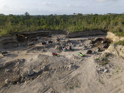 Aerial view of an active excavation site with heavy machinery digging pale earth, sharply contrasting with the lush green forest, Klaten, Central Java, Indonesia.