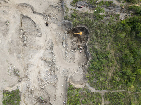 Aerial view of an excavator digging into the pale, scarred earth, sharply contrasting with the vibrant, untouched green foliage on the right, Klaten, Central Java, Indonesia.