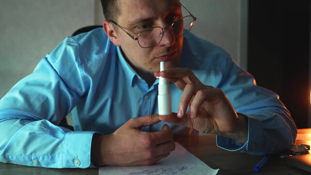 Businessman suffering from stuffy nose using nasal spray to reduce swelling and ease breathing. Concept of working while sick in evening office.