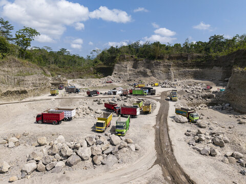 Aerial view of an expansive open-pit quarry bustling with vibrant trucks moving light-toned rocks and earth under a vast blue sky, Klaten, Central Java, Indonesia.