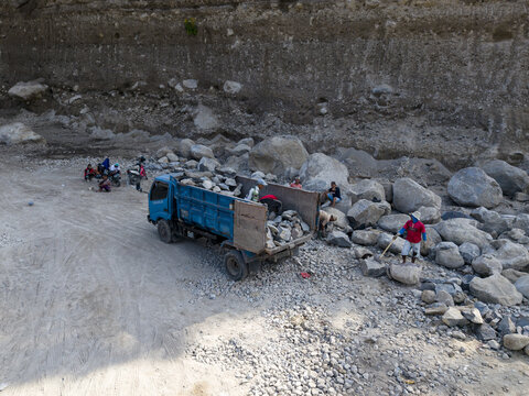 Aerial view of strenuous labor as workers load immense rocks onto a blue truck amidst rugged terrain and a towering dirt wall, Klaten, Central Java, Indonesia.