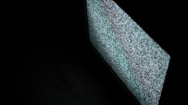 Close angled shot of a television screen showing flickering analog static noise pattern against a dark background, representing no signal or broadcast interference. Technology concept footage.