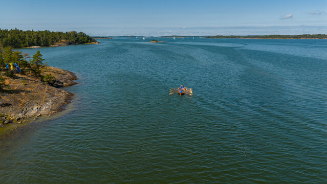 Aerial view of a small, colorful raft gliding through deep green waters, contrasting with the rugged, tree-lined coast under a clear sky, Hiittinen, Southwest Finland, Finland.