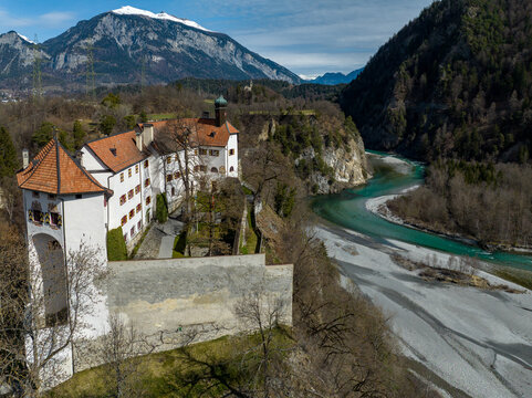 Aerial view of the imposing white-walled Rhazuns Castle perched above the vibrant turquoise river, framed by rugged mountains under a clear sky, Rhazuns, Grisons, Switzerland.