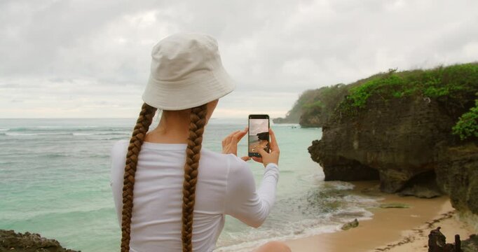 Woman capturing serene beach cove with phone during quiet tropical morning