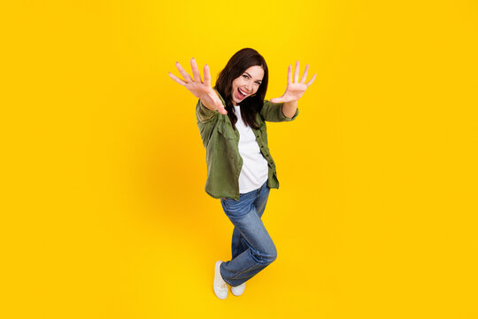 Happy young woman reaching out and laughing on a bright yellow background in casual jeans and green shirt, energetic playful pose