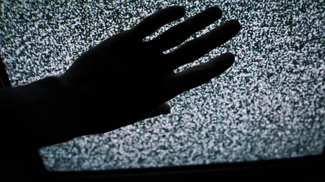 Silhouette of a hand reaches out and touches a television screen displaying flickering static noise. The mysterious interaction suggests paranormal activity or fear.