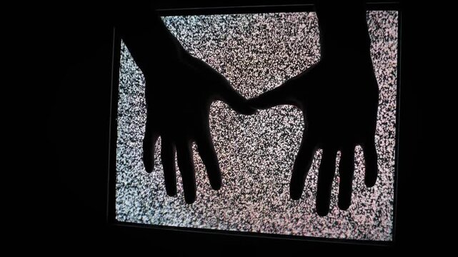 Two dark hand silhouettes descend slowly towards an old CRT television screen displaying flickering analog static noise. Spooky horror concept, poltergeist theme, suggesting interaction from beyond.