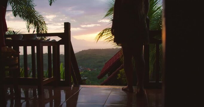 Barefoot woman stands on a balcony watching the colorful sunset over the valley