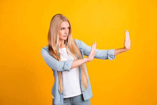 Young woman making stop gesture and setting a boundary while pushing away with hands on bright yellow background, casual denim shirt and serious confident expression