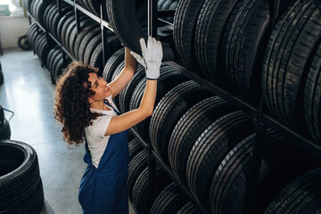 Female auto technician with curly hair wearing gloves organizes tires on a shelf in a tire storage...