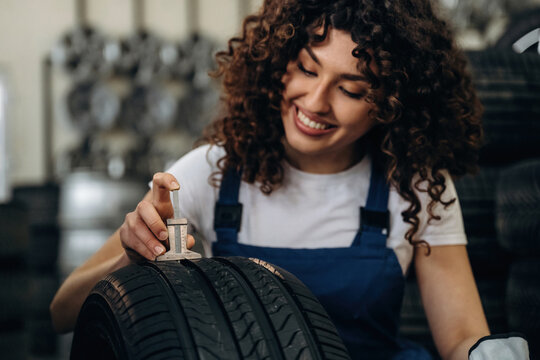 Female mechanic with curly hair inspects tire tread depth using a gauge in a tire shop, stacks of tires visible in the background