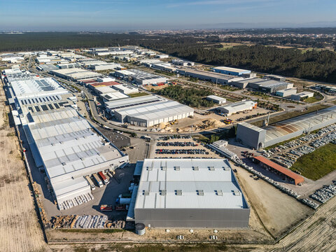 Aerial view of a sprawling industrial zone with gleaming grey warehouses under a bright sky, bordered by dark green forests, Vagos, Aveiro, Portugal.