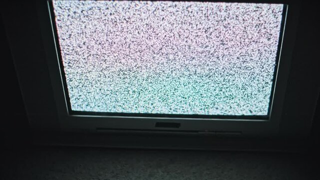 Close view from above of static noise (snow) filling a vintage CRT television screen. The old monitor displays white and colored pixelation in a dimly lit room.