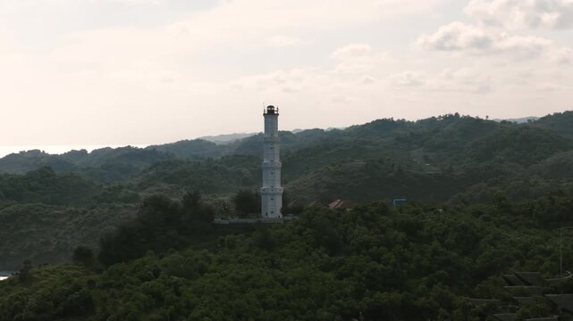 Aerial view of Mesra Beach lighthouse standing on a lush green hill overlooking the coastline under a cloudy sky in Kecamatan Tanjungsari, Daerah Istimewa Yogya, Indonesia.