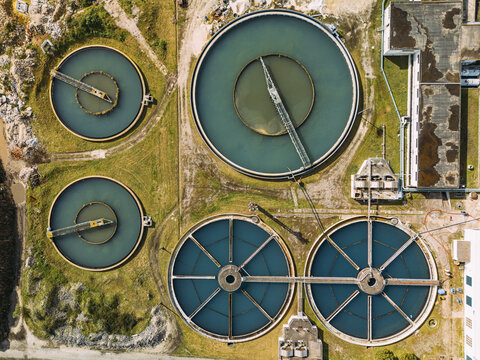 Aerial view of a vast water treatment plant, where stark concrete structures frame circular basins swirling with deep blues and murky greens under a clear sky, Cacia, Aveiro District, Portugal.